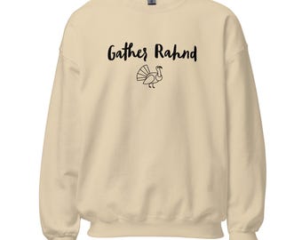 May include: A cream-colored sweatshirt with the words "Gather Rahnd" in black script above a simple turkey illustration. The long-sleeved crewneck top is made of a soft material.