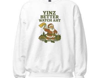 May include: White sweatshirt with the text "YINZ BETTER WATCH AHT" in green. The graphic features Santa Claus in a red suit, waving a yellow flag, with a city skyline and Christmas trees.