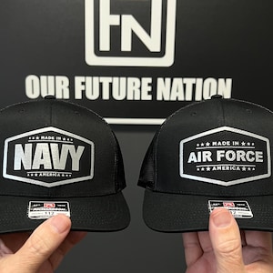 Puede incluir: Dos gorras de camionero negras con paneles traseros de malla. Una gorra presenta un parche plateado con el texto "***MADE IN*** NAVY ***AMERICA***" y la otra gorra presenta un parche plateado con el texto "***MADE IN*** AIR FORCE ***AMERICA***".