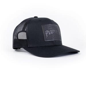 May include: Black trucker hat with a black embroidered patch that says "Blue Collar Brewery" and "Plumber" in a vintage font.