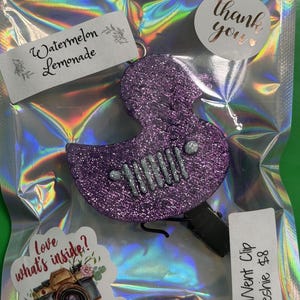 May include: A purple glittery duck-shaped air freshener with a silver seven-slot grille design on the front. The item is packaged in a holographic plastic bag with labels reading Watermelon Lemonade, love what's inside?, and Small Vent Clip Freshie.