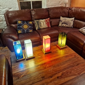 May include: Four stained glass lamps on a wooden table. The lamps are in shades of blue, white, yellow, and red. The lamps are on a wooden table in front of a brown leather couch.