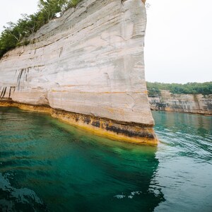 May include: A tall, layered rock cliff with green trees at the top, rising out of clear, turquoise water. The cliff is a light beige color with darker brown streaks.