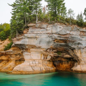 May include: A rocky cliff with a cave opening, covered in green trees, overlooking a turquoise blue lake.