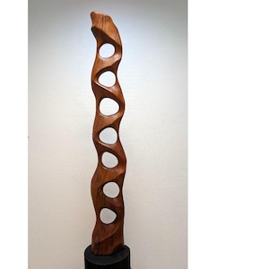 Può includere: Una scultura in legno alta e verticale caratterizzata da una tonalità marrone calda e venature naturali. L'oggetto presenta una forma ondulata con una serie di sei fori circolari che attraversano il centro, creando una struttura aperta.