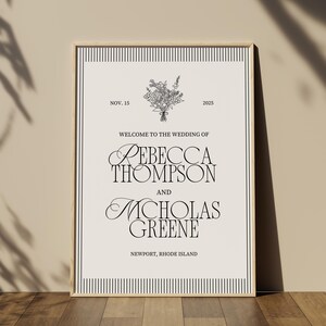 May include: A framed wedding welcome sign with a cream background and black text. The sign includes the names Rebecca Thompson and Nicholas Greene, the date November 15, 2025, and the location Newport, Rhode Island. A floral illustration is at the top.