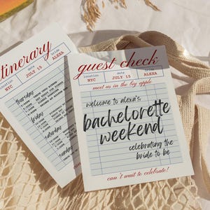 May include: Two white paper cards with red and black text. One card reads "Itinerary" and the other "Guest Check". Both cards have the date "July 15" and the name "Alexa". The "Guest Check" card says "welcome to alexa's bachelorette weekend".