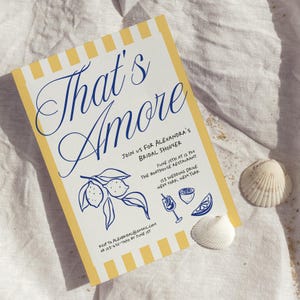 May include: A bridal shower invitation with the words "That's Amore" in blue script. The invitation has a yellow and white striped border and features illustrations of lemons and cocktails. Two seashells are placed near the invitation.