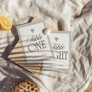 May include: Two white table number cards with black text and a striped border. The cards read "Table ONE" and "Table EIGHT" with the names "Rebecca & Nicholas" below. A seashell and a half lemon are also visible.