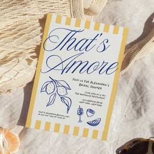 May include: A bridal shower invitation with a yellow and white striped border. The invitation features the words "That's Amore" in elegant blue script. Additional text includes details for the event, along with lemon illustrations.