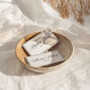 May include: Two place cards with names and table numbers rest in a speckled ceramic bowl. The cards have a white background with black text and a thin black border. The top card reads "Rachel Green, Table One." The bottom card reads "Alexa Milton, Table Two."