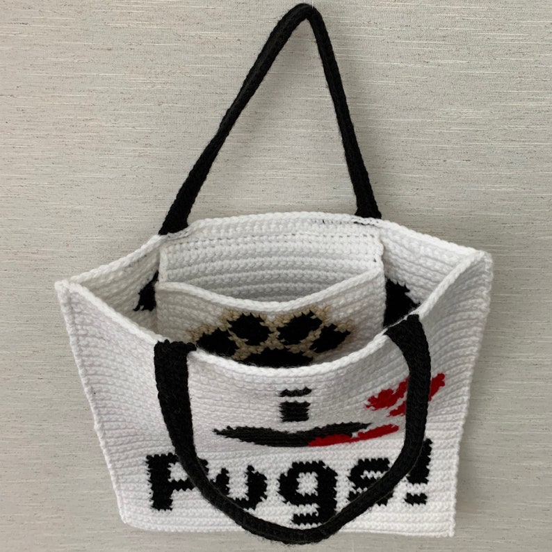May include: White crocheted tote bag with black handles and a paw print design. The bag has the word "Pugs!" written on the front in black.