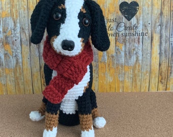 May include: A handmade crochet dog with black, white, and brown details, wearing a red scarf. The dog is sitting upright, with a black nose and large eyes. The background is a wooden surface with the text "just to Create own sunshine" and "Crochet Pattern only."