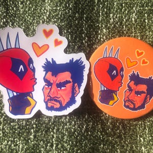 Könnte beinhalten: Ein gestanzter Aufkleber und ein runder Ansteckbutton mit einer Cartoon-Illustration einer rot maskierten Figur mit drei silbernen Klingen und einer dunkelhaarigen Figur mit Bart. Drei orangefarbene Herzen befinden sich zwischen den Figuren.