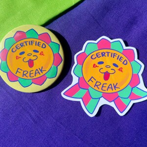 May include: A circular pin-back button and a die-cut sticker, both featuring a yellow sun-like character with a pink and green scalloped border. The design includes the text "CERTIFIED FREAK" in blue, with a smiling face showing a tongue.