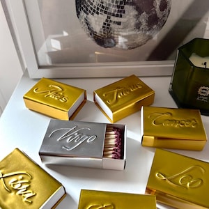 Puede incluir: Una colección de cajas de cerillas rectangulares con signos del zodiaco en relieve en acabados metálicos dorados y plateados. Las cajas están etiquetadas como Aries, Taurus, Cancer, Virgo, Leo, Libra y Gemini. Una caja Virgo plateada está abierta, revelando cerillas de madera con puntas de color morado oscuro.