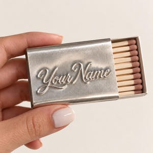 May include: A rectangular silver-toned metal matchbox case featuring the embossed script text Your Name on the front surface. The case is partially open, revealing a row of wooden matches with light brown tips arranged inside the white inner tray.