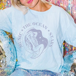 May include: A light blue t-shirt featuring a graphic of a mermaid within a circle, with the text "Music * The Ocean * Stars". The model is wearing the shirt with a light blue headband and is seated against a sparkly iridescent backdrop.