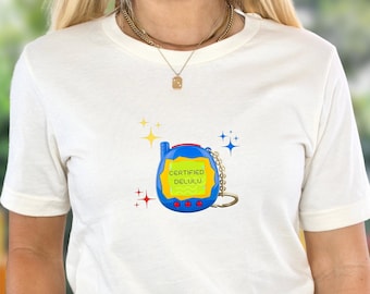 May include: White t-shirt with a blue and yellow cartoon image of a digital pet with the text "CERTIFIED DELULU" on the screen.