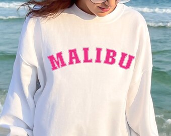 Op de afbeelding: Witte sweatshirt met het woord "MALIBU" in felroze letters op de borst. De sweatshirt heeft lange mouwen en een ronde hals. De achtergrond toont een strand en de zee.