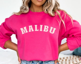 Op de afbeelding: Een felroze sweatshirt met het woord "MALIBU" in een crèmekleurig lettertype in collegestijl over de borst. De sweatshirt heeft lange mouwen en een ronde hals. Het model draagt een blauwe spijkerbroek.