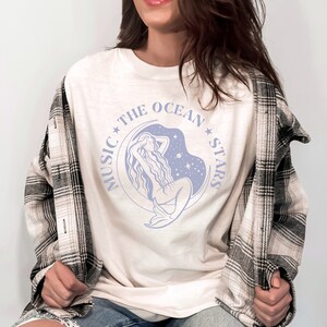 May include: White t-shirt with a blue graphic of a mermaid sitting on a crescent moon with stars and the text "Music The Ocean Stars".