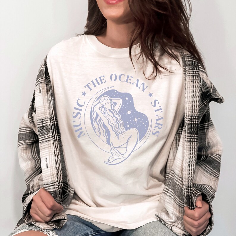 May include: White t-shirt with a blue graphic of a mermaid sitting on a crescent moon with stars and the text "Music The Ocean Stars".