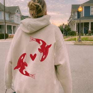 May include: A beige hoodie with a red orca design and a heart. The orcas are facing each other. The hoodie has a hood and long sleeves. The background shows a residential street with houses and a cloudy sky.