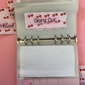 May include: A pink leather bound planner with a clear plastic pocket on the front. The pocket contains a cherry patterned sticker that says "Going Out".
