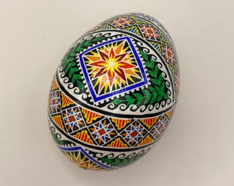 Puede incluir: Un huevo pysanka pintado a mano con intrincados patrones geométricos en tonos de rojo, naranja, amarillo, verde, azul y negro. El huevo está decorado con un diseño central en forma de estrella y un borde de hojas estilizadas.