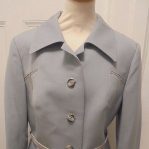 Può includere: Una giacca di tailleur blu chiaro con una cintura bianca e quattro bottoni argentati. La giacca ha un colletto a revers e maniche lunghe.