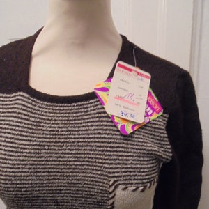 Könnte beinhalten: Ein brauner und weißer gestreifter Pullover mit einem braunen Strickkragen und kurzen Ärmeln. Der Pullover hat einen Patch auf der Vorderseite und ein Preisschild, das an der Schulter befestigt ist. Das Preisschild lautet "ARTIKEL 181, GRÖSSE 38, FARBE 151, 100% Synthetics, 34.50".