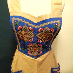 Könnte beinhalten: Eine beige Schürze mit blauem Oberteil und Taschen. Die blauen Bereiche sind mit floralen Mustern in Rot, Gold und Grün bestickt. Die Schürze hat breite Träger und ein tailliertes Oberteil im Vintage- oder Folk-Stil.