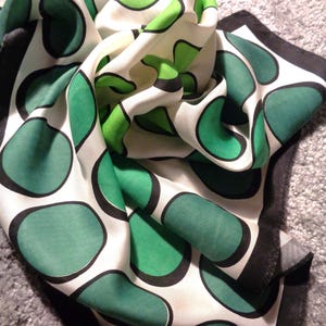 Puede incluir: Un pañuelo cuadrado con fondo blanco y grandes formas redondeadas de color verde delineadas en negro. El pañuelo tiene un borde negro sólido a lo largo de los extremos y está drapeado en pliegues, mostrando una textura suave y brillante.