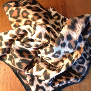 Puede incluir: Un pañuelo cuadrado con un estampado de leopardo clásico que presenta manchas negras, marrones y tostadas sobre un fondo claro. La tela tiene un acabado suave y ligeramente brillante, rematado con un borde fino cosido en color negro.