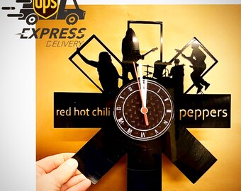 May include: Black vinyl record clock featuring a band silhouette design with the words "red hot chili peppers". The clock has a round face with Roman numerals and red hands. The clock is mounted on a gold background.