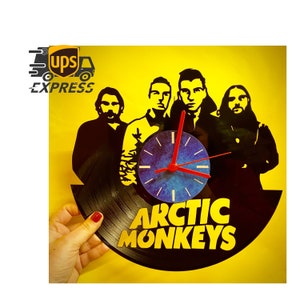 May include: A black vinyl record clock featuring the band Arctic Monkeys. The clock face is blue with red hands, and the band members are silhouetted above the band's name. The background is yellow.