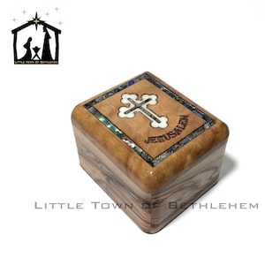 May include: A small wooden box with a cross and the word "Jerusalem" inlaid on the top. The box is made of olive wood and has a natural wood finish. It measures 6.80 inches long, 6.00 inches wide, and 4.70 inches high.