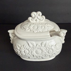 May include: White ceramic soup tureen with a lid and a spoon slot. The tureen features embossed floral designs and decorative handles. The lid is topped with a scrolled finial. The tureen is set against a dark background.