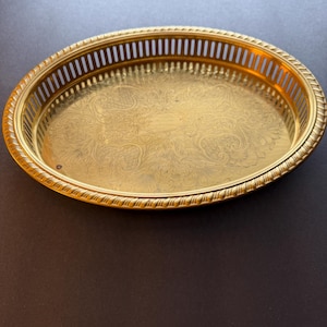 May include: A gold-colored, oval serving tray with a detailed floral pattern in the center. The tray has a decorative rope-like edge and a slotted border. The tray is made of metal and is suitable for serving food or displaying items.