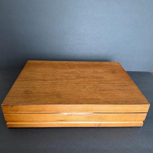 May include: A rectangular wooden box with a light brown finish. The box has a lid and a recessed handle. The wood grain is visible on the surface. The box is sitting on a dark surface.