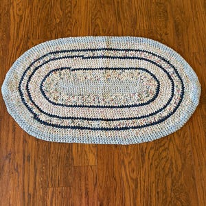 May include: An oval-shaped, handmade rug with a light blue and multi-colored speckled pattern. The rug features two navy blue concentric oval borders. The rug is made of a soft, textured material and is suitable for a bathroom or entryway.