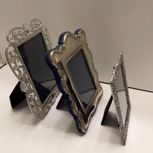 May include: Three ornate silver picture frames of varying sizes. The frames have decorative edges and dark blue or black backing. Each frame is propped up with a black stand, ready for display. The frames are on a white surface.