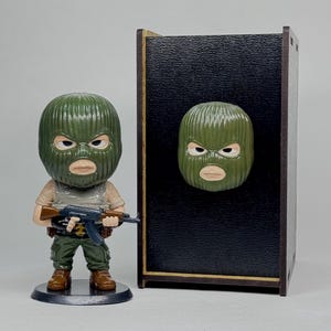 May include: A figurine of a person wearing a green ski mask and holding a rifle. The figurine is standing next to a black box with a matching mask attached. The figurine is wearing a tan shirt, green pants, and brown boots.