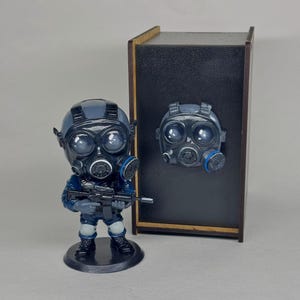 May include: A figurine of a soldier in a gas mask and blue uniform, holding a rifle. A gas mask is attached to a black box. The figurine is on a black base. The gas mask has blue accents.