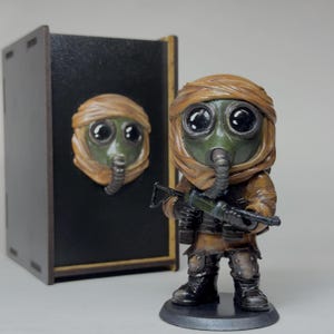 May include: A detailed figurine of a figure in a gas mask and military attire, holding a rifle. The figure is brown and green, with large, expressive eyes. A matching gas mask is mounted on a black box.