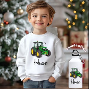 Peut inclure: Un sweat-shirt blanc avec un tracteur vert et jaune et le nom "Hugo" en écriture noire. Une gourde assortie est également présente. L'arrière-plan comprend un sapin de Noël et des lumières festives.