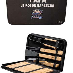 Op de afbeelding: Zwarte barbecuegereedschapsset met de tekst "PAPA LE ROI DU BARBECUE" en een grillafbeelding. De set bevat een spatel, tang, vork, mes en snijplanken. De gereedschappen hebben houten handvatten en worden opgeborgen in een etui met ritssluiting.