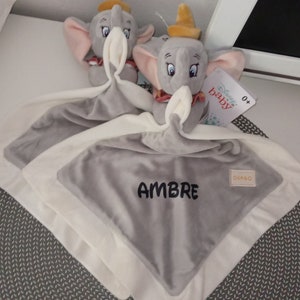 Peut inclure: Une couverture bébé grise et blanche avec un jouet en peluche d'éléphant gris. La couverture est brodée du nom "AMBRE". Le jouet en peluche d'éléphant a un chapeau jaune et tient une étiquette qui dit "Disney Baby".