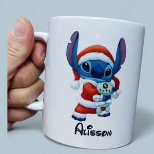 K&ouml;nnte beinhalten: Wei&szlig;e Keramiktasse mit einem blauen Cartoon-Charakter in einem Weihnachtsmann-Outfit, der ein kleines Pl&uuml;schtier h&auml;lt. Auf der Tasse steht der Name "Alisson" in schwarzer Schrift. Die Tasse hat einen wei&szlig;en Henkel.